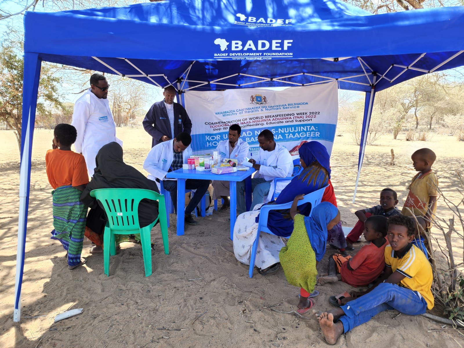 Health workers in white coats receive families under a BADEF tent at the free mobile clinic in Qorax IDP camp, Dhobley