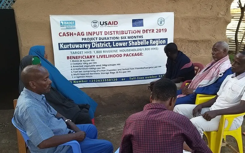 Beneficiaries seated beside a BADEF cash and agricultural input distribution banner in Kurtunwarey